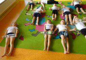 Dzieci leżą na plecach, butelki plastikowe turlają po brzuchu i nogach.